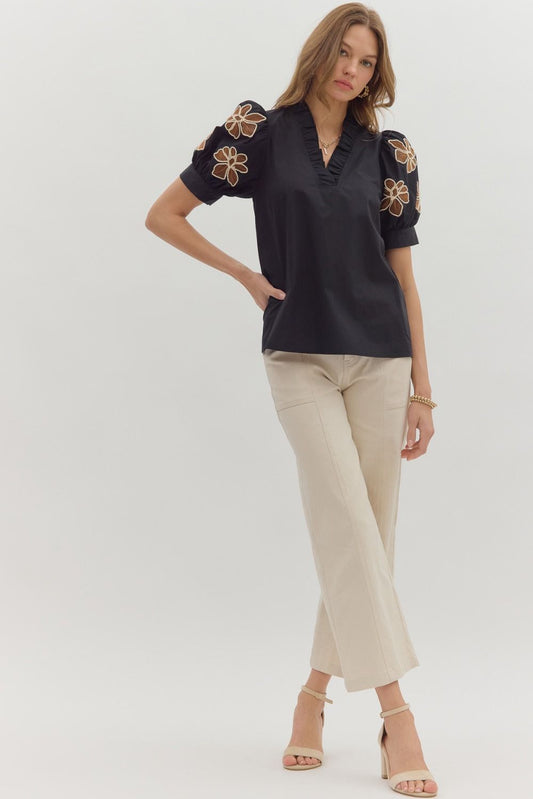 Woman wearing a black blouse with floral embroidery and beige pants on a white background