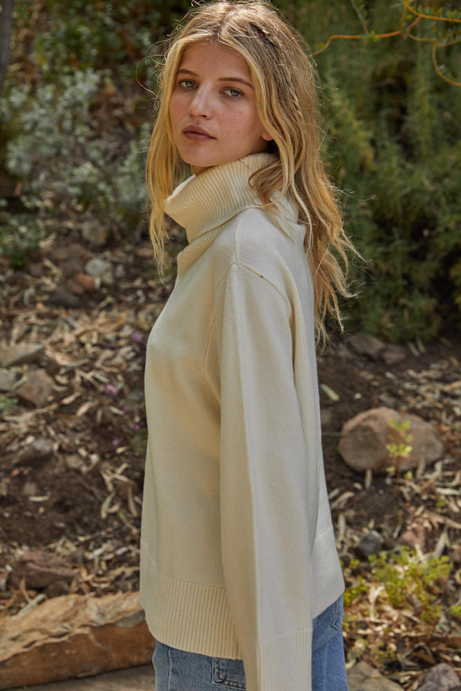 Woman wearing a beige sweater in a natural setting with greenery.