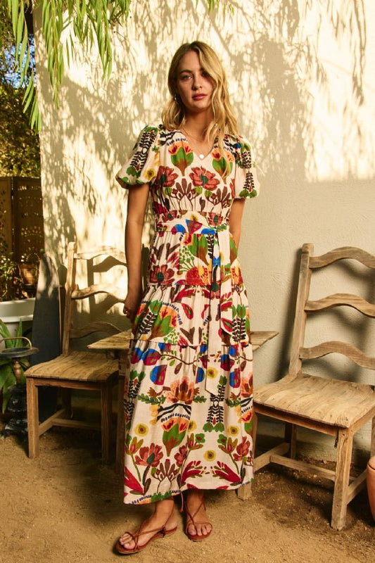 Woman in a colorful floral dress standing in a sunlit outdoor setting with wooden chairs.